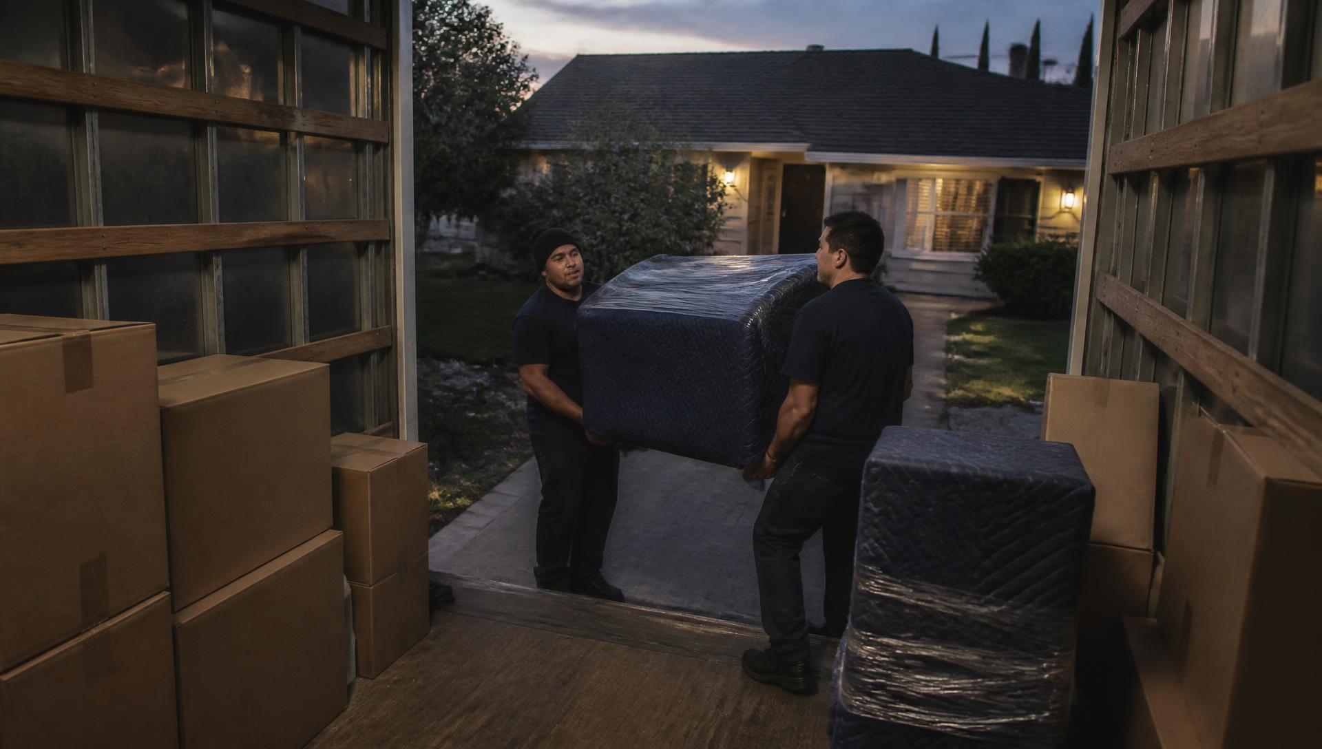 A moving crew loading blanket-wrapped furniture into a residential moving truck at dusk
